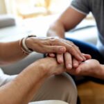 Man holding senior woman's hand at home. Male healthcare worker holding hands of senior woman at care home, focus on hands. Shot of a young man hands holding old senior elderly hand with love and care